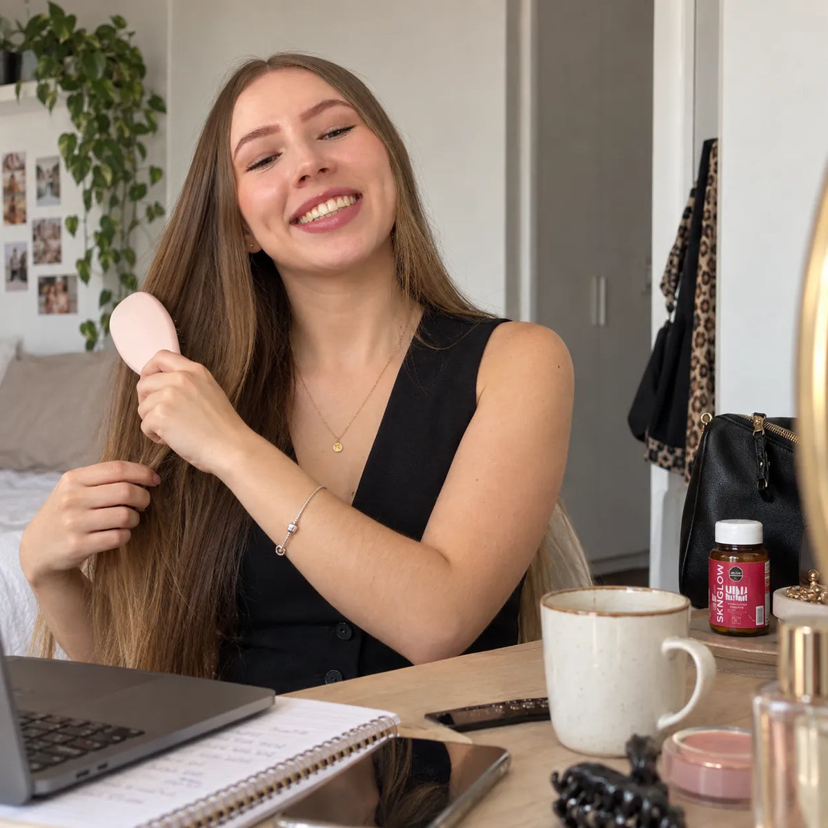 Mujer sonriente se cepilla el cabello largo en su escritorio; a su lado, una taza y un frasco de Sknglow